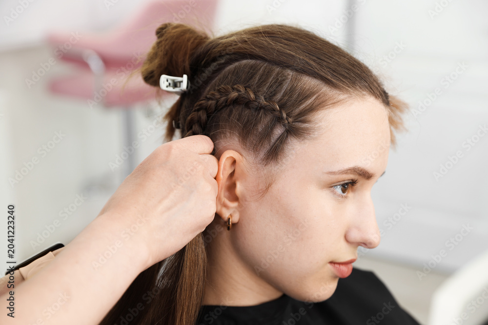 Fototapeta premium Hairstylist braiding teenage girl's hair in salon, closeup