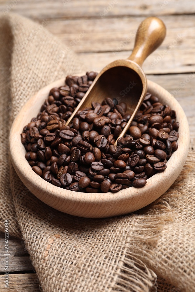 Fototapeta premium Coffee beans in bowl, scoop and burlap fabric on wooden table, closeup
