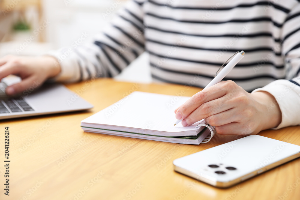 Fototapeta premium Woman taking notes while working with laptop at wooden table indoors, closeup