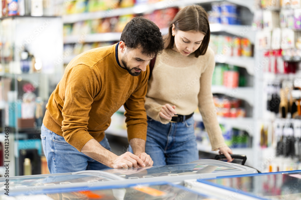 Fototapeta premium Positive young couple choosing cooled food product standing at the fridge in grocery store