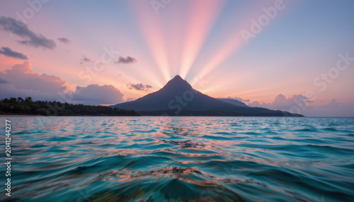 Sunset rays spread behind mountain over calm ocean water with distant tropical island and soft clouds in sky