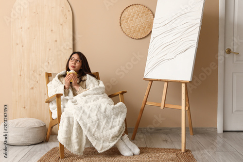 Young happy Asian woman with cup of cacao and plaid sitting on armchair near ...