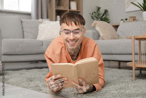 Tableau sur toile Young tattooed man in eyeglasses reading book on floor at home