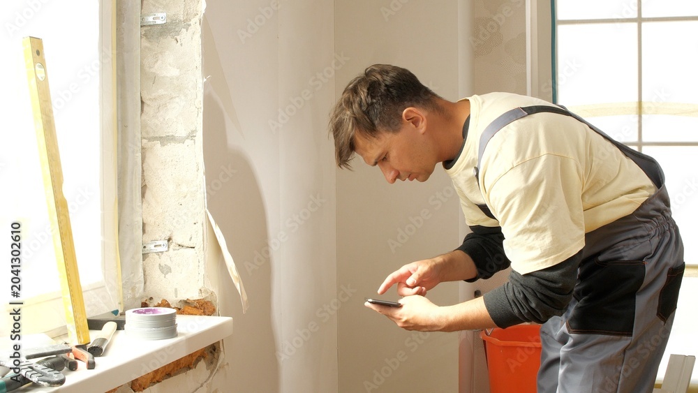 Fototapeta premium Construction worker in overalls using a smartphone, checking details or communicating during a home renovation project, standing by a window with construction tools on the sill