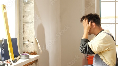 Quadro su tela Man in work clothes sitting near a partially torn down wall, covering his face w