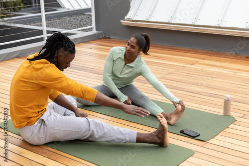 Diverse couple in activewear stretching on rooftop deck with green mats, water bottle, smartphone