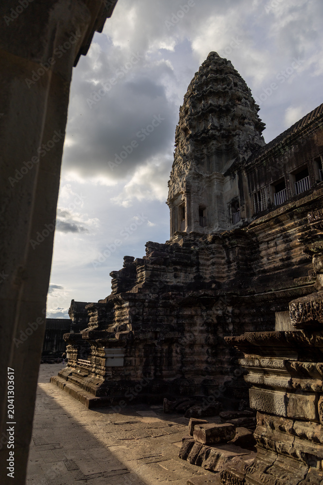 Fototapeta premium Angkor Wat temple complex in Siem Reap, Cambodia, at sunset. The ancient Khmer architecture is bathed in dramatic light and shadow under a cloudy sky.