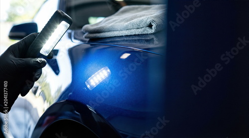 Car detailing professional inspecting blue vehicle paint with LED light in automotive workshop, surface quality control and paint correction concept.
