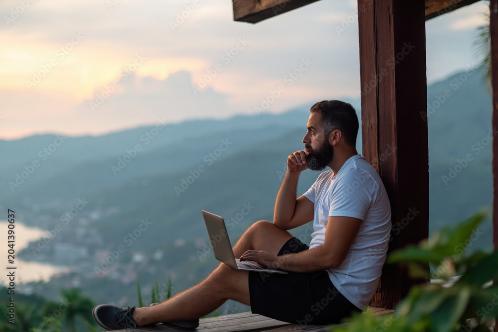 Obraz premium Man bearded using laptop on balcony with mountain view at sunset remote work