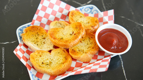 close-up crispy toasted garlic bread slices on red checkered paper served with ketchup on black marble table.