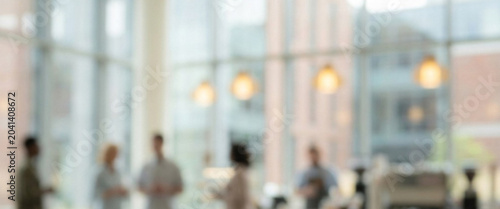 A blurred image capturing a modern indoor space with large windows, featuring groups of people interacting in a casual environment, suggesting a social or professional setting.