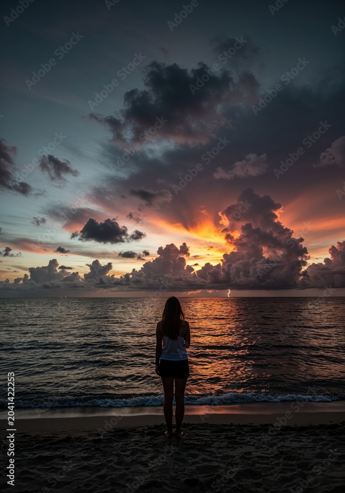 Fototapeta premium A Moment of Solitude at Dusk: A solitary figure stands on a sandy beach, gazing out at the tranquil ocean as the sky ignites with the fiery hues of sunset.