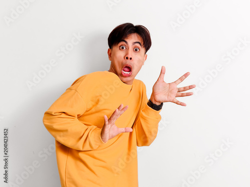 Canvastavla A surprised young Asian man in a yellow sweatshirt stares directly ahead with a funny wide-eyed shocked expression