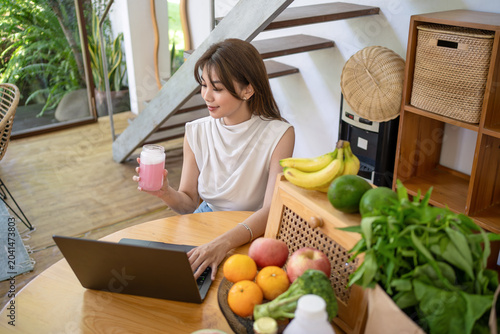 Asian Woman Drinking Healthy Smoothie While Working on Laptop at Home
