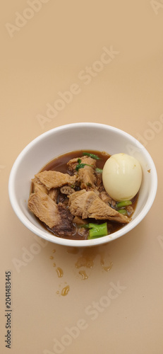Braised pork leg with hard boiled egg in brown soup, Thai food known as Kha Moo Phalo, served in a white bowl on cream background.