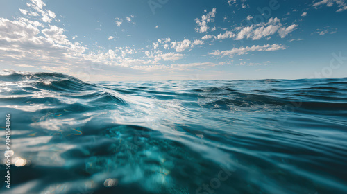 Close-up view of clear blue ocean water with sunlight creating ripples and reflections on the surface, under a sky with scattered clouds.