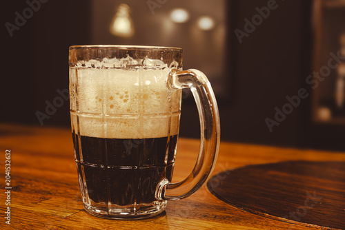 Foto Dark stout beer in a mug with white foam on wooden bar counter