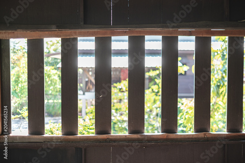 View through wooden slats of a traditional house, revealing greenery and sunlight outside.