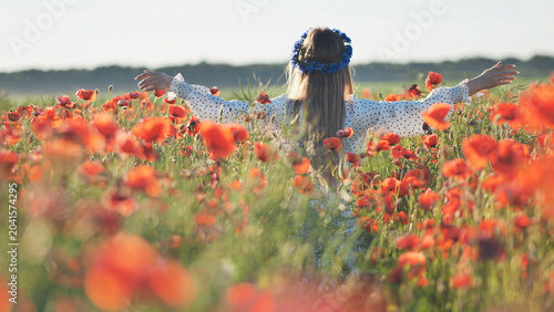 Young woman with long blond hair, wearing a blue flower crown and white dress, embracing the beauty of a vibrant poppy field