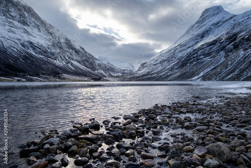 A beautiful cloudy day at a mountain lake with stone pebbles on the shore. A winter scenery on Eidsvatnet in Norway, Geiranger area, UNESCO world heritage site.