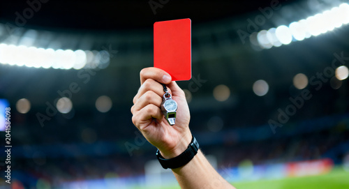 Referee's hand holds a red card and whistle, ready to signal a foul or expulsion during a professional sports match. Authority and game rules enforcement in action