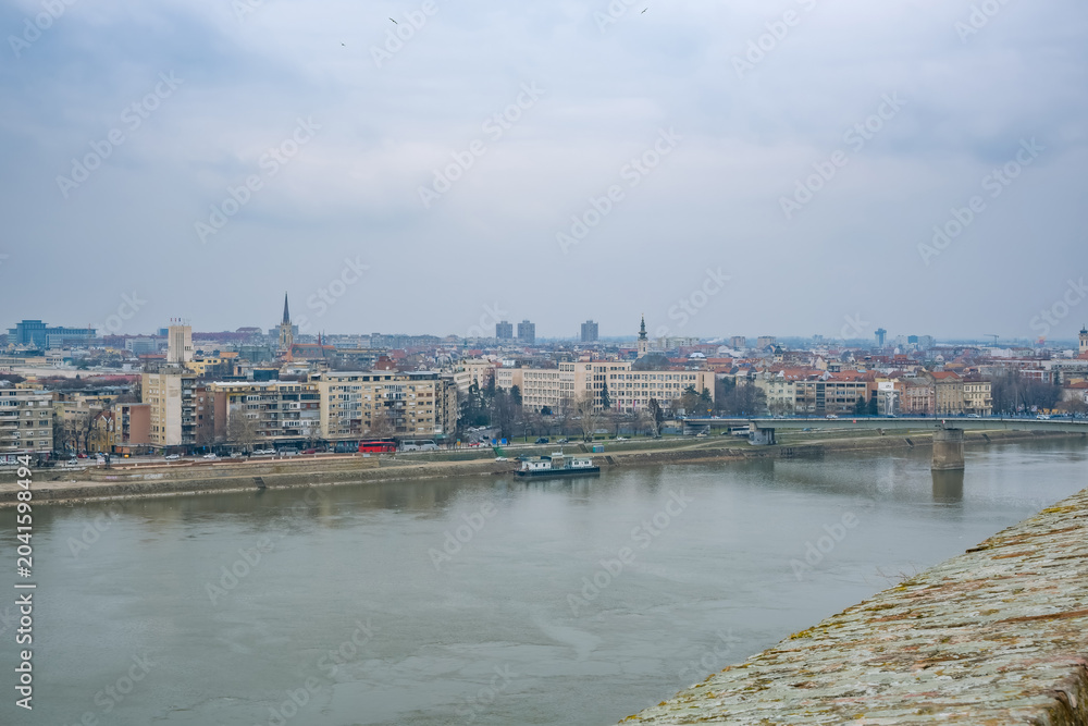 Obraz premium A panoramic view of Novi Sad, Serbia, showcasing the city skyline along the Danube River. Buildings line the riverbank under a cloudy sky.