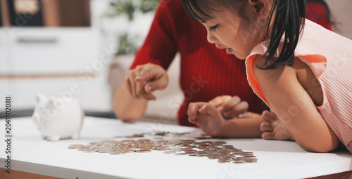 A mother and her little daughter. The happy schoolgirl, a kindergarten child, is smiling and counting coins into her piggy bank. Parents and child are saving money. Financial literacy at home.