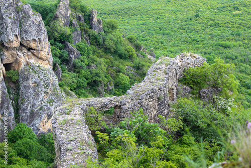 A stone fortress wall on a cliff covered with grass and bushes. Grass and forest in the background. Khornabuji, Kakheti, Georgia
