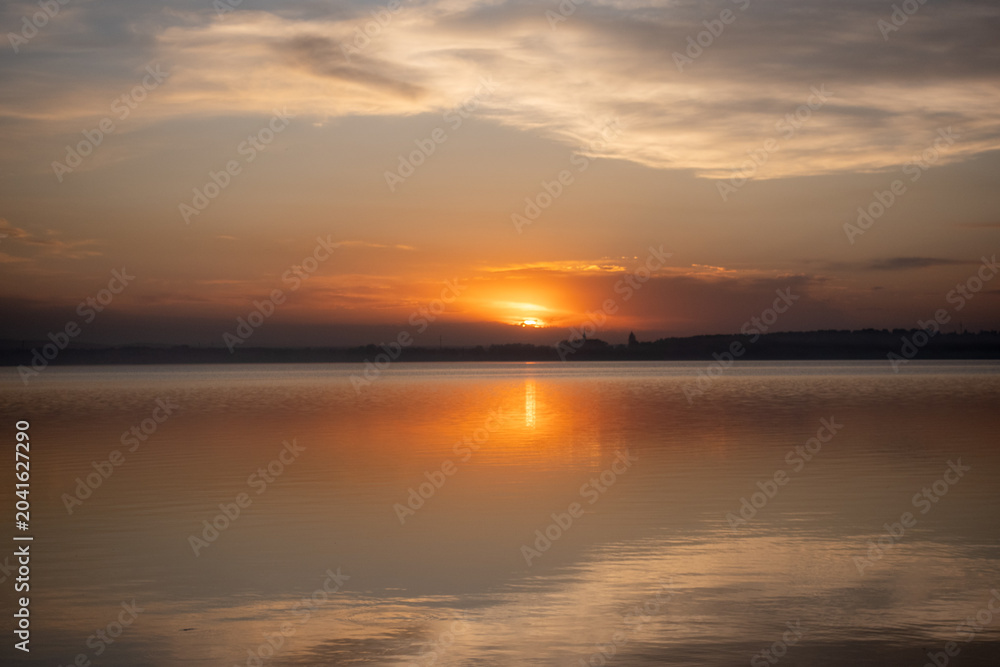Fototapeta premium Serene sunset over a calm lake with reflections and distant silhouette of buildings
