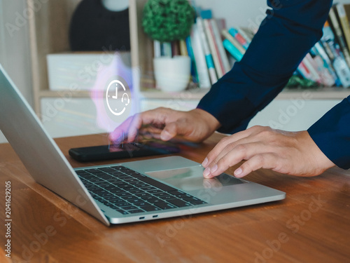 Hands typing on laptop keyboard with smartphone nearby and holographic clock icon symbolizing productivity and time management in home office setting