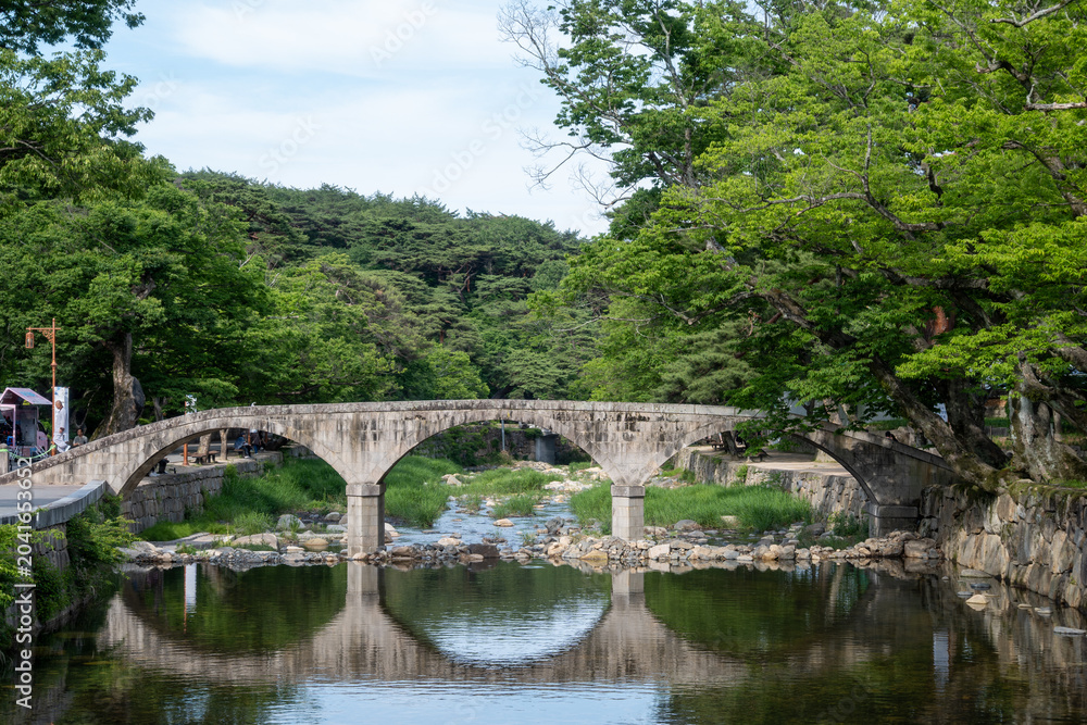 Fototapeta premium Traditional Korean arched stone bridge reflecting perfectly on the calm water of a stream surrounded by lush green trees