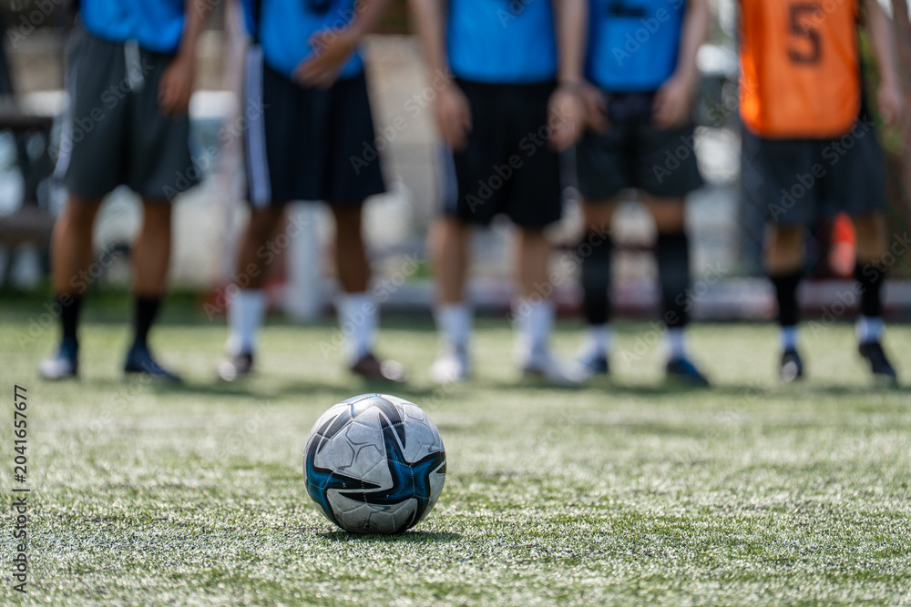 Naklejka premium Close up of soccer ball on green artificial turf pitch with blurred players background, football match training session concept before kickoff at sports stadium