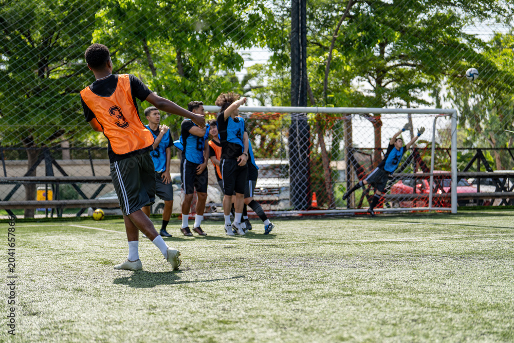 Naklejka premium African American soccer player taking free kick shot during football match. Diverse opponents defensive wall standing near goal post waiting for ball outdoor