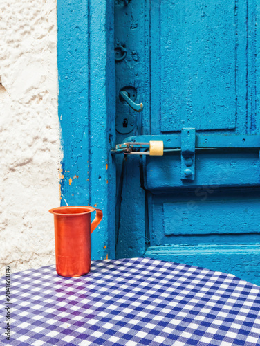 Rustic outdoor cafe setting in Athens, Greece, featuring a blue checkered tablecloth, copper cup, and textured blue door.