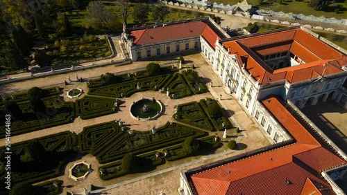 Queluz National Palace showing its intricate French style formal gardens with hedges and fountains, capturing the historic architecture from an aerial perspective under bright daylight
