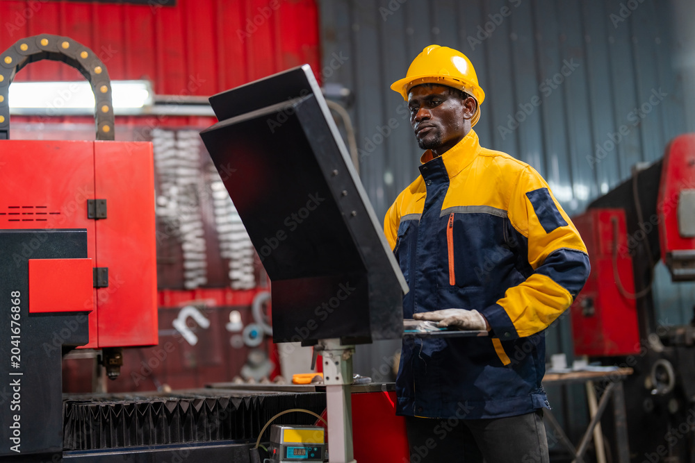 © VStudio - Male worker wears a yellow hard hat and blue-yellow safety jacket. He stands near a black monitor in a factory setting. Red industrial machinery is visible behind him. Focused work environment