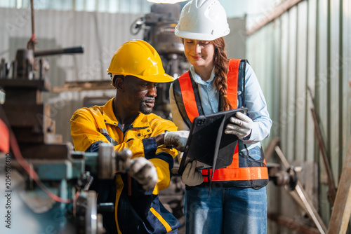Man in a yellow hard hat and safety vest interacts with machinery. Woman in a white hard hat and orange vest holds a tablet. They discuss data together in an industrial factory setting