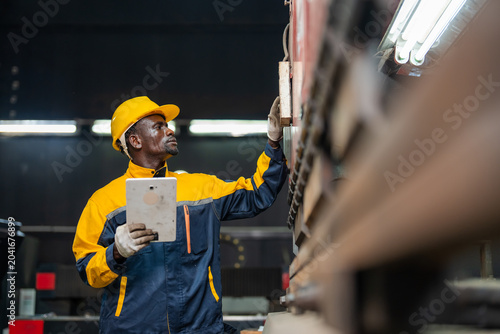 Worker in yellow hard hat and blue jacket holds clipboard. He inspects a large metal structure. Industrial setting with bright overhead lighting. Focus on his hands and the machinery