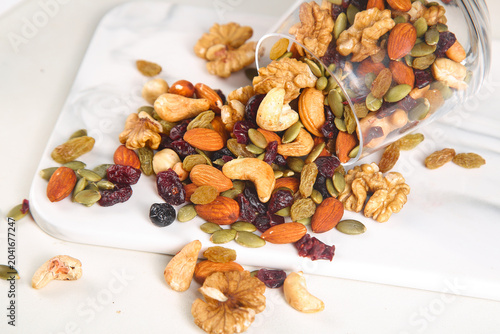 Assorted Nuts and Dried Fruits Trail Mix Spilling from Glass Jar on White Wood Background