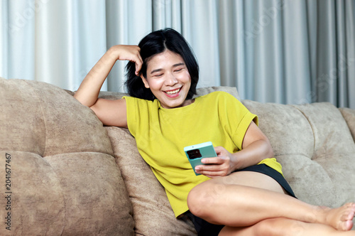 Happy young Asian woman using smartphone on sofa at home, representing lifestyle, social media, online communication, entertainment, and positive emotions