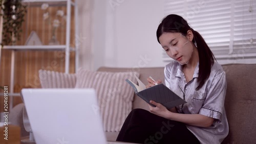 young Asian woman read notebook write journal morning routine relax sitting couch laptop home living room peaceful calm lifestyle activity start day plan