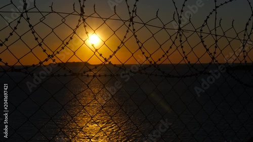 Border at sunset. Orange sun setting over calm body of water, seen through a metal fence topped with barbed wire, creating a poignant contrast between natural beauty and confinement