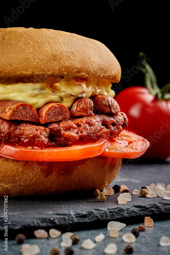 Gourmet vegan burger with plant-based meat patty, sausage slices, sauce and fresh tomatoes on black slate board, side view of delicious alternative protein burger on dark background