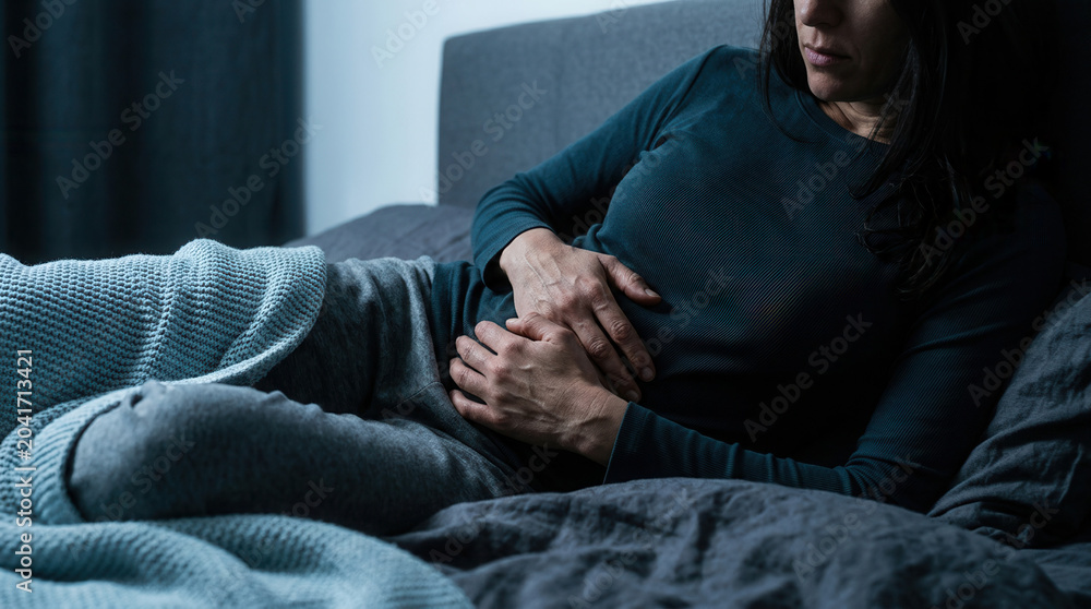 Fototapeta premium Woman Lying in Bed Clutching Her Abdomen Feeling Discomfort in Dark Lighting