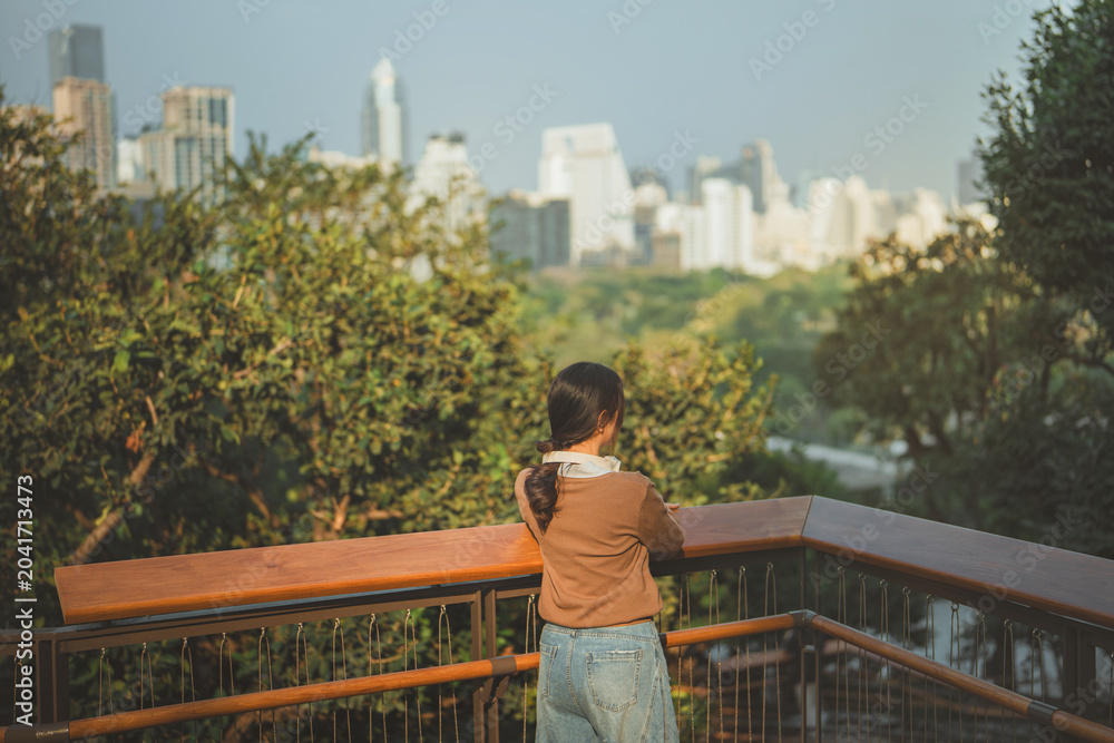 Naklejka premium Rear view of young Asian woman leaning on wooden balcony rail and looking at city skyscrapers, thoughtful female relaxing at observation deck park in morning, modern urban life, mindfulness and future