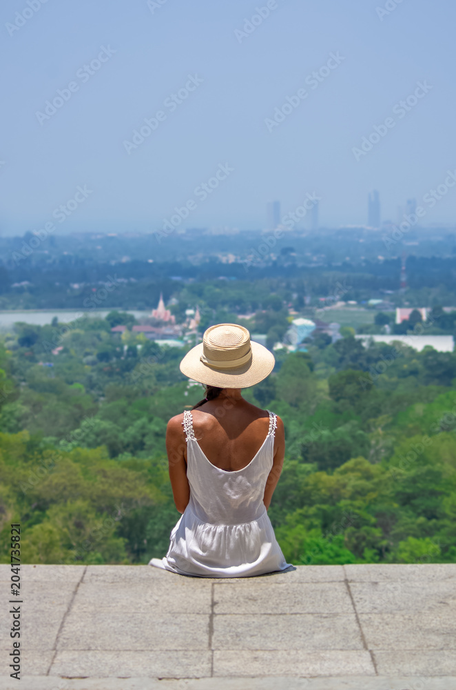 Fototapeta premium Woman in white dress and straw hat sitting on hilltop viewpoint overlooking tropical city landscape in Thailand