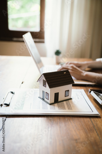 A miniature house placed on a contract document while a professional works on a laptop, symbolizing real estate transactions, property management, home financing, and digital business solutions.