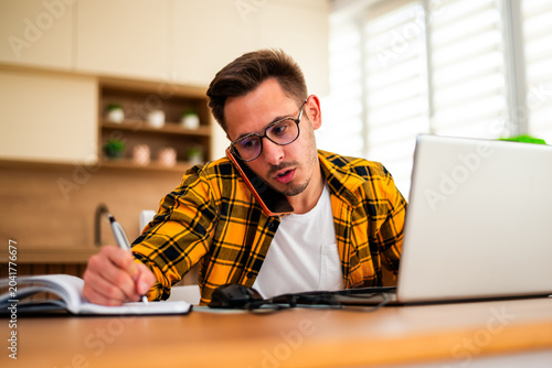 Working from home, a young man pauses to write down ideas and reminders, combining online research with traditional note-taking.