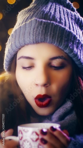 Woman blows the hot drink in hands surrounded by Christmas lights