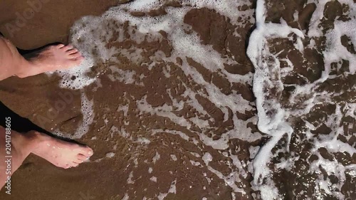 first person point view woman feet standing on sandy shore while gentle sea waves with white foam wash over them, evoking sense calm and relaxation during summer vacation.